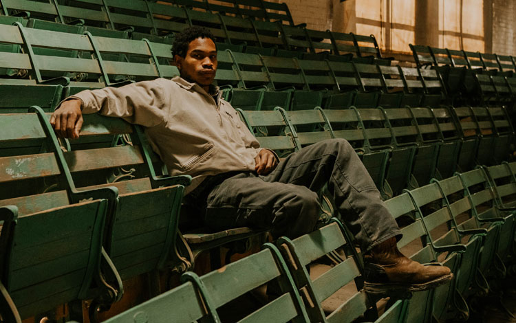 A man sits in the stands at the cattle sale barn, posing and wearing workwear.